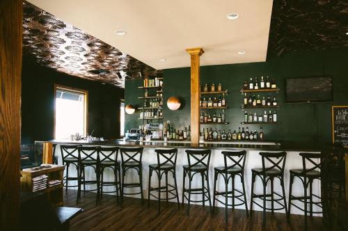 A stylish bar with a wooden counter, high stools, and shelves lined with various bottles against a dark green wall.
