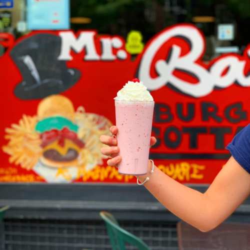 A person holds a large strawberry milkshake topped with whipped cream and a cherry, with a colorful restaurant mural in the background.