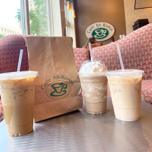 Three iced coffee drinks and a brown paper bag on a table at Café Ah-Roma, with a cozy seating area in the background.