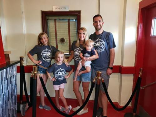 A family of four in matching gray shirts poses in a movie theater lobby with a ticket counter in the background.