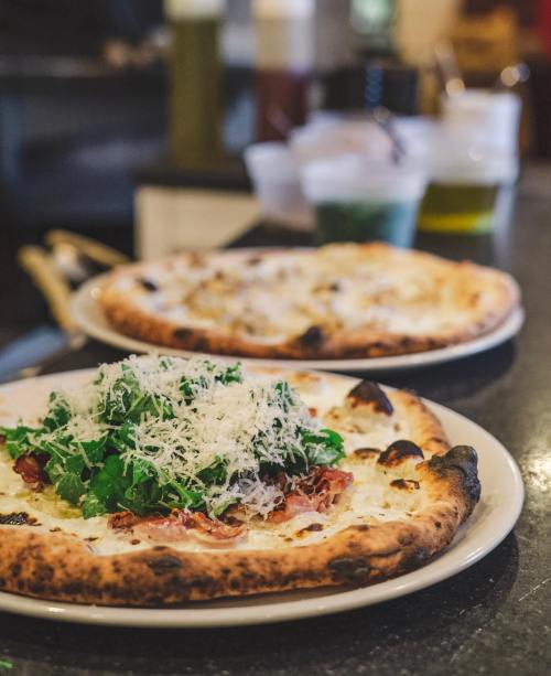 A close-up of a pizza topped with greens and cheese, with another pizza in the background on a dark countertop.