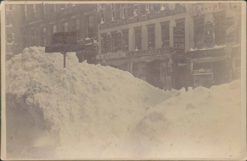 A snowy street scene with large snowdrifts and old buildings in the background, creating a winter atmosphere.