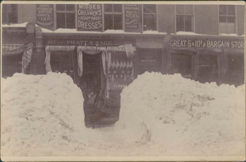 Historic snowy street scene with a storefront displaying dresses and a bargain store sign. Snow piles on either side.