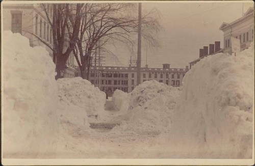 Snow-covered street with large snowbanks on either side, lined by bare trees and historic buildings in the background.