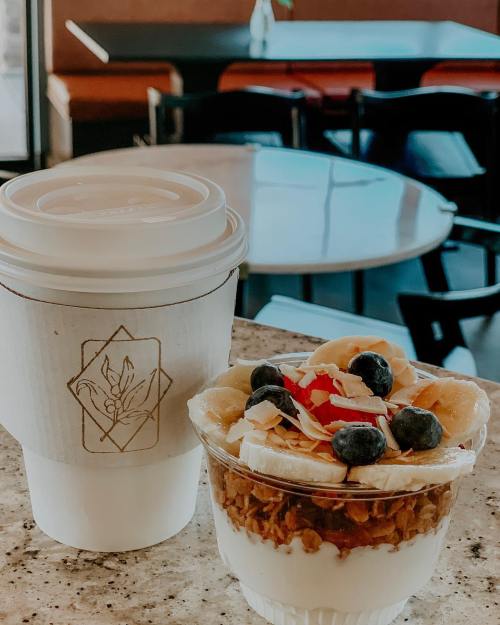 A coffee cup beside a parfait with granola, yogurt, banana, blueberries, and coconut flakes on a table.