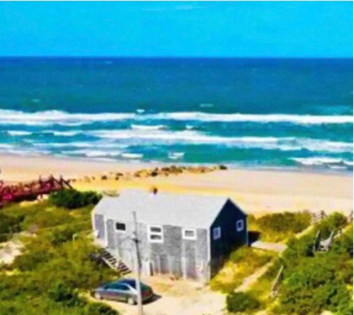 A small gray beach house surrounded by greenery, overlooking a sandy beach and ocean waves under a clear blue sky.