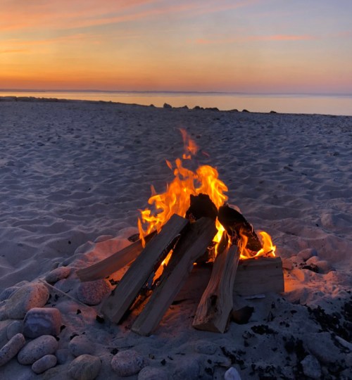 A cozy campfire on a sandy beach at sunset, with calm waters and a colorful sky in the background.