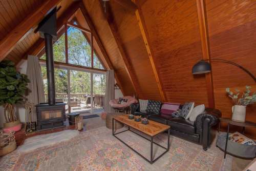 Cozy A-frame living room with wooden walls, a fireplace, and large windows overlooking nature. Stylish decor and furniture.
