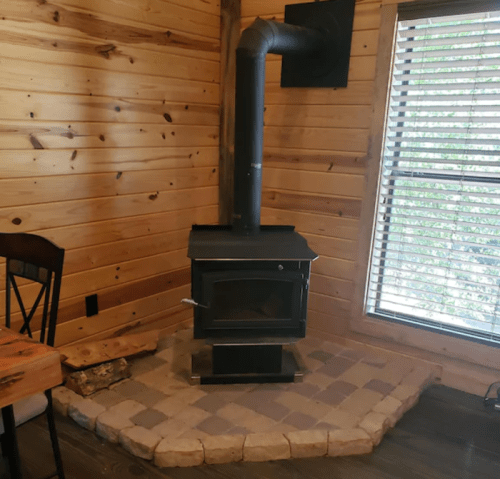 A black wood stove in a wooden cabin, with a stone base and a vent pipe, beside a window with wooden blinds.