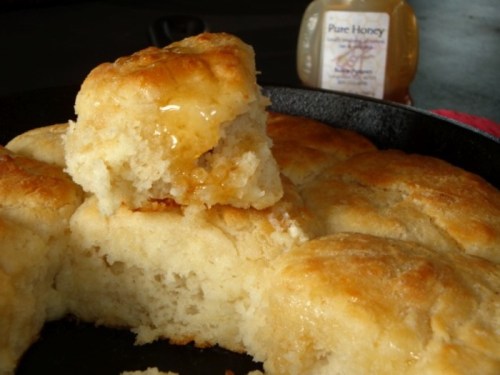 Golden, fluffy biscuits in a cast iron skillet, with one biscuit topped with honey, and a jar of honey in the background.