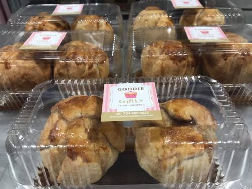 Clear plastic containers filled with freshly baked cinnamon rolls, each labeled with "Goodie Girls Cake Shoppe."