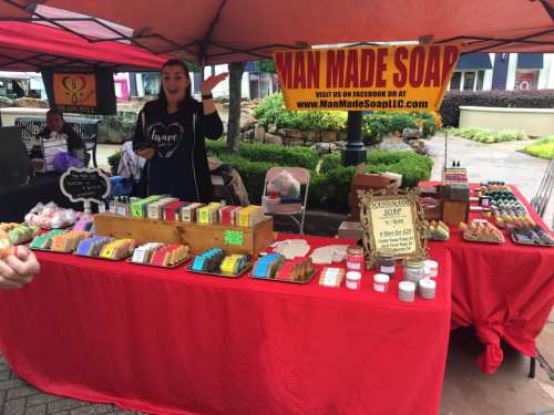 A vendor at a market booth showcasing colorful handmade soaps and greeting customers under a red tent.