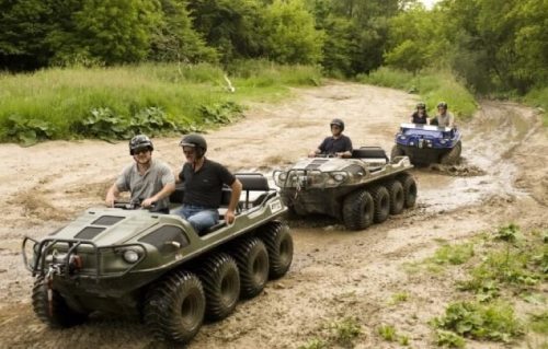 Four people ride all-terrain vehicles on a muddy trail surrounded by greenery.