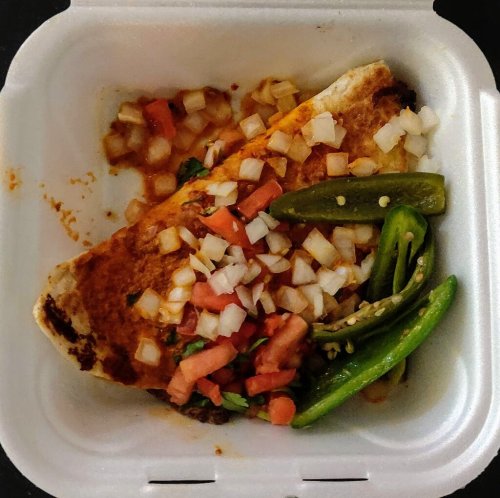 A takeout container with a burrito topped with diced tomatoes and onions, alongside green peppers.