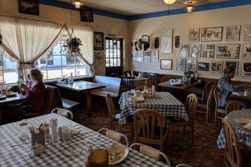 A cozy diner with checkered tablecloths, empty tables, and two patrons enjoying their meals by the window.