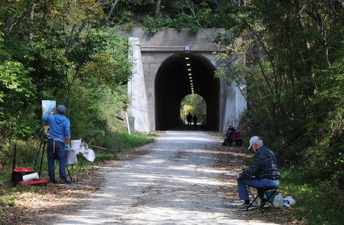 Two artists paint along a path near a tunnel, with trees surrounding the area and two figures walking in the distance.