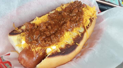 A loaded hot dog topped with chili and shredded cheese, served in a toasted bun on a paper-lined tray.