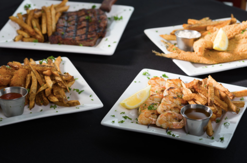 Four plates of food featuring steak, fried shrimp, and crispy fries, garnished with lemon and herbs on a black tablecloth.