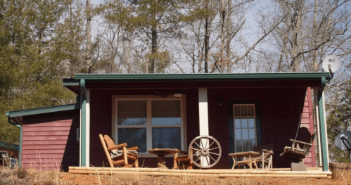 A cozy red cabin surrounded by trees, featuring a porch with rustic chairs and a wagon wheel decoration.
