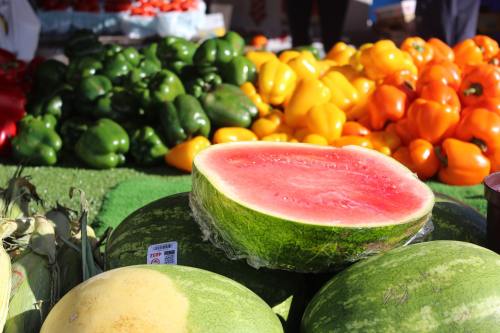 A cut watermelon sits atop whole watermelons, with colorful bell peppers and other produce in the background.