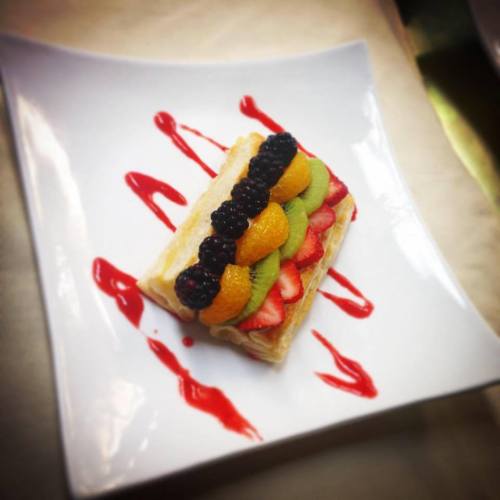 A layered pastry topped with strawberries, kiwi, oranges, and blackberries, drizzled with red sauce on a white plate.