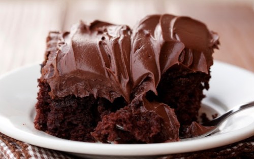 A slice of rich chocolate cake with smooth chocolate frosting on a white plate, set on a wooden table.