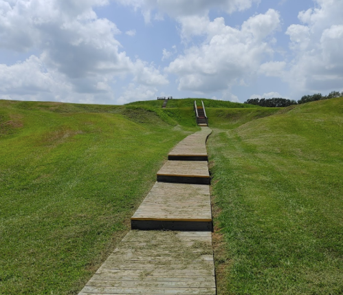 A wooden pathway leads up a grassy hill under a partly cloudy sky. Steps are visible at the top of the hill.