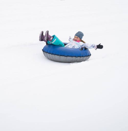 A child in winter clothing joyfully sledding down a snowy hill on a blue and gray inner tube.