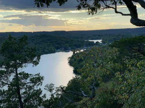 A serene view of a lake surrounded by trees, with a sunset casting golden light over the water and hills.