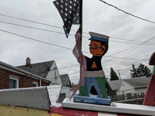 A painted wooden figure of a sailor holding an American flag, set against a cloudy sky and rooftops in the background.