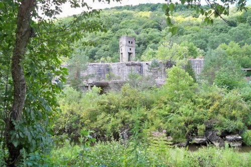 Abandoned structure surrounded by lush greenery and trees, with a tower visible against a hillside backdrop.