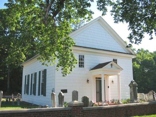 A white, historic building with black shutters, surrounded by trees and a cemetery, under a clear blue sky.