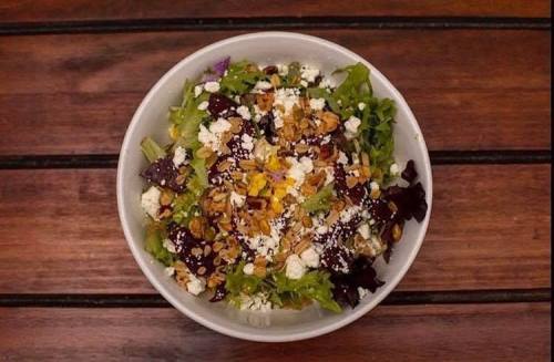 A bowl of mixed greens topped with crumbled cheese, nuts, and colorful vegetables on a wooden table.