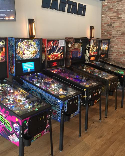 A row of colorful pinball machines in a brick-walled arcade, featuring various game designs and vibrant lights.