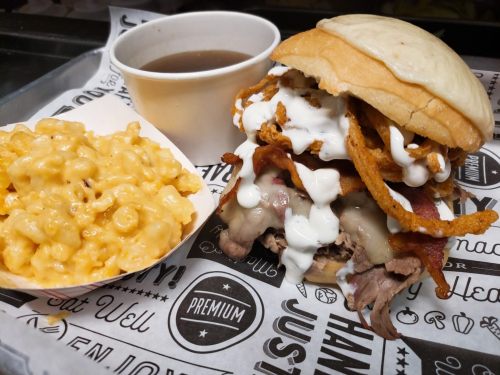 A hearty sandwich with beef, bacon, and toppings, served with macaroni and cheese and a cup of sauce on the side.