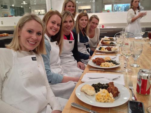 A group of women in aprons sit at a table with plates of food, smiling and enjoying a cooking class together.