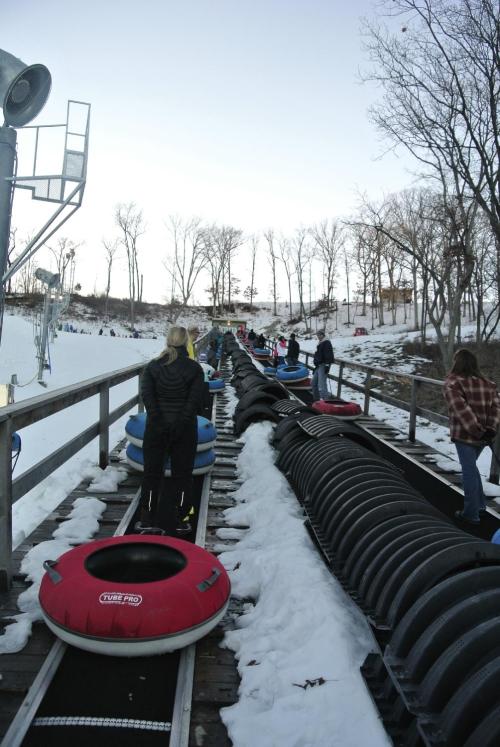 A snowy hill with people waiting in line for tubing, surrounded by trees and a clear sky.