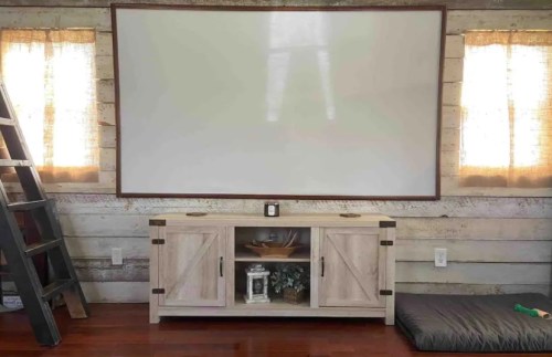 A cozy room with a large blank screen, wooden cabinet, and a ladder against a rustic wall, featuring soft curtains.