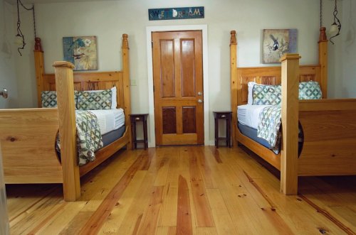 A cozy bedroom with two wooden beds, decorative pillows, and a wooden door, featuring warm wooden floors.