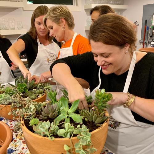 A group of women in aprons planting succulents in terracotta pots during a workshop.