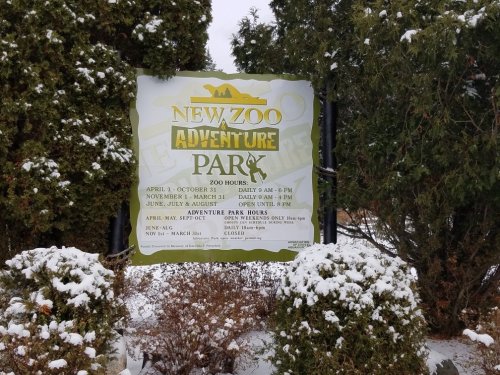 Sign for New Zoo Adventure Park with hours and seasonal details, surrounded by snow and greenery.