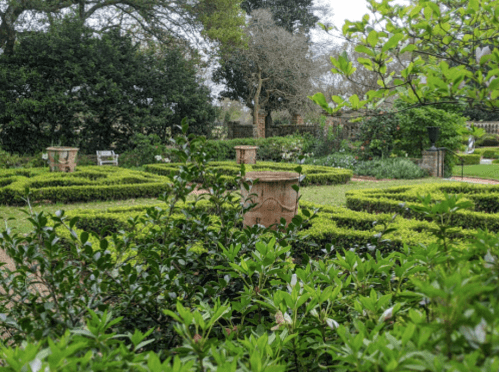 Lush garden with neatly trimmed hedges, stone planters, and benches surrounded by greenery.