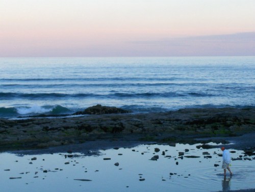 A person walks along a rocky beach at sunset, with gentle waves and a calm sea reflecting the pastel sky.