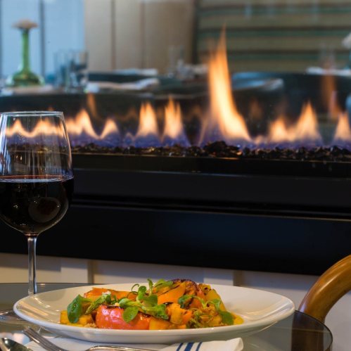 A plate of colorful food sits in front of a glass of red wine, with a cozy fireplace glowing in the background.