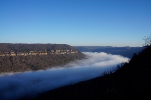A scenic view of a valley with fog rolling through, surrounded by steep cliffs and a clear blue sky.