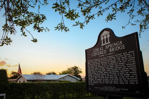 Sign for Coral Springs Covered Bridge with a sunset sky and trees in the background. American flag visible.