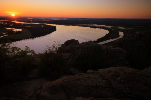 Sunset over a winding river, with rocky foreground and lush green landscape, casting warm hues across the sky.