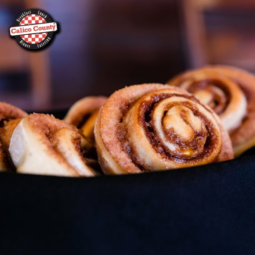 Freshly baked cinnamon rolls with swirls of cinnamon and sugar, served in a black basket. Calico County logo in corner.