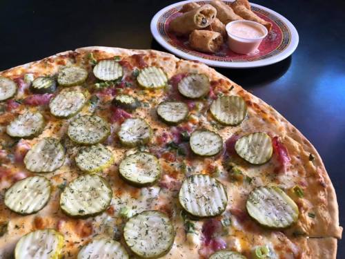 A large pizza topped with pickles and herbs, alongside a plate of spring rolls and dipping sauce.