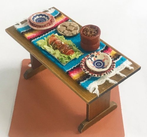 A miniature table set with colorful textiles, featuring plates of food, including lettuce and small bowls of snacks.